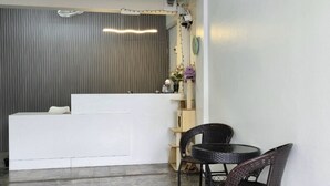 Een receptie met een wit bureau, rieten stoelen, een tafeltje en een decoratieve plank met planten.