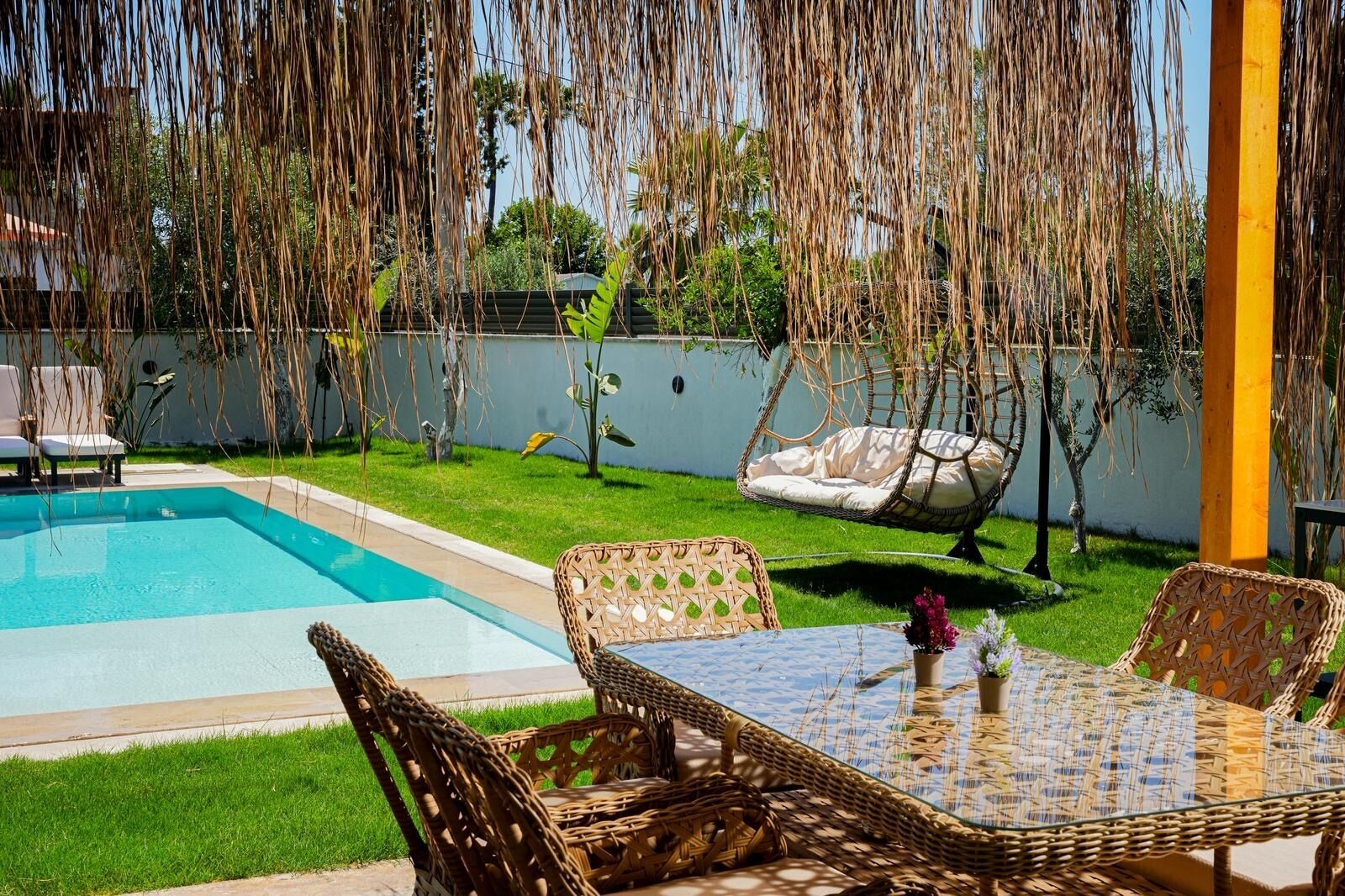 A poolside area with a wicker table and chairs, a hanging chair, and a small garden with a fence.
