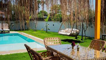 A poolside area with a wicker table and chairs, a hanging chair, and a small garden with a fence.
