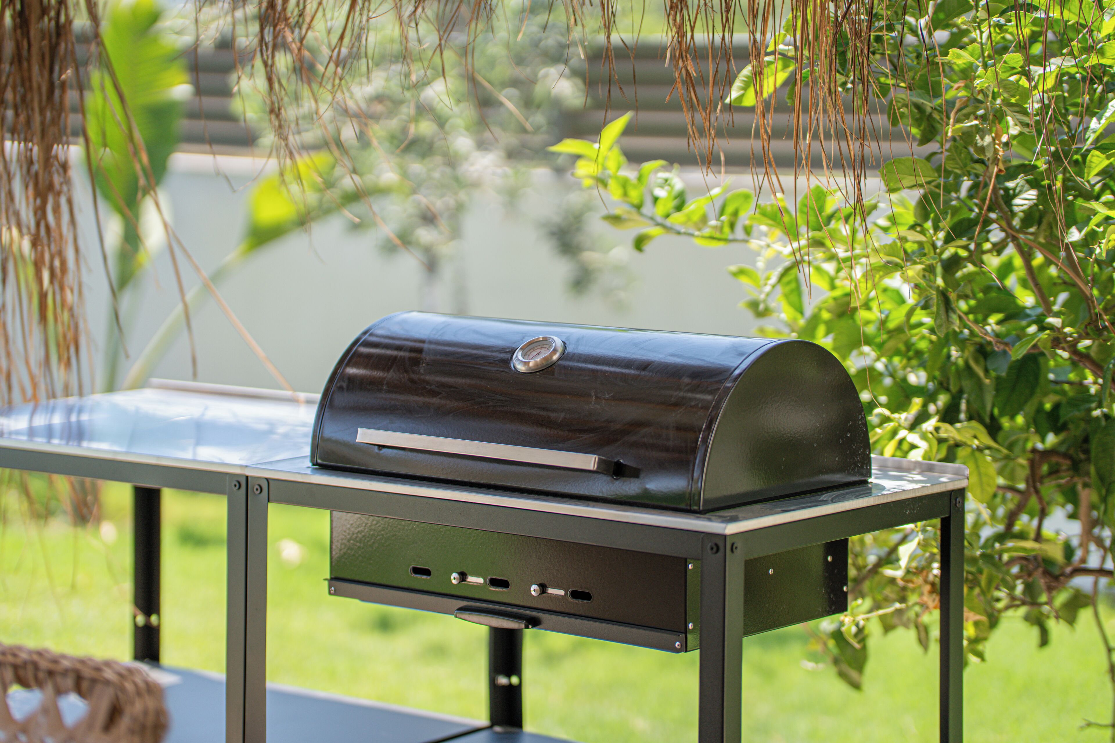 A stainless steel barbecue grill on an outdoor table with a metal frame, surrounded by greenery.