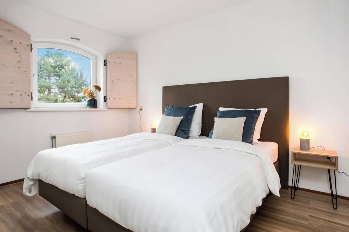 Een tweepersoonsbed met witte beddengoed en twee kussens, een nachtkastje met een lamp, en een raam met uitzicht op bomen.