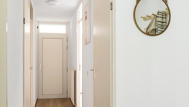 A hallway with wooden flooring, white walls, and doors. A round mirror hangs on the wall. - Beachside Home With Balcony (Julianadorp)