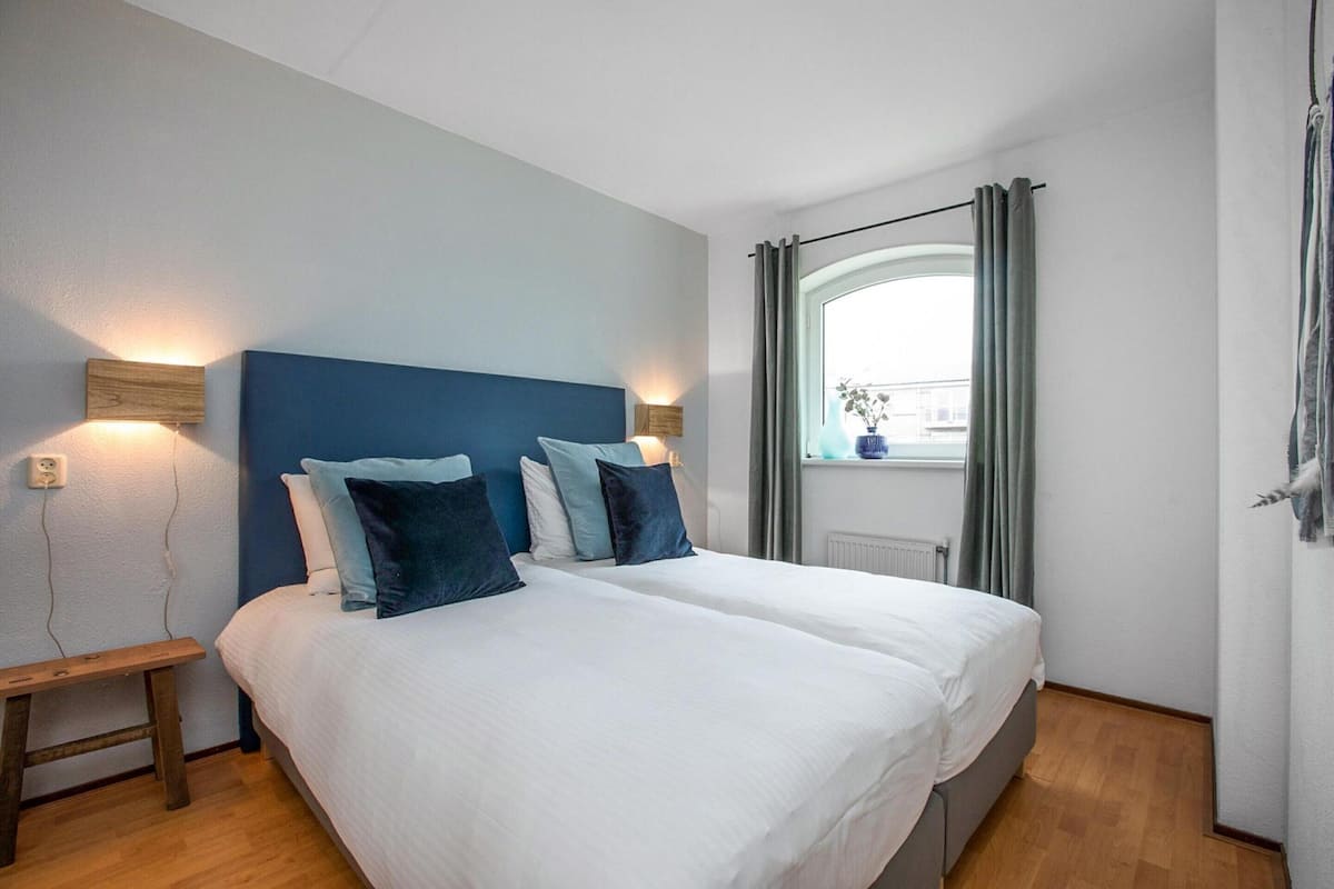 Een tweepersoonsbed met witte beddengoed en blauwe kussens, een nachtkastje en een raam met gordijnen.