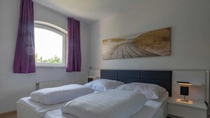 A double bed with white linens, a headboard, a bedside table with a lamp, and a window with purple curtains. - Beachside Home With Balcony (Julianadorp)