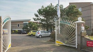 A gated entrance with a brick building, a sign indicating a speed limit of 15, and parked cars. - Beachside Home With Balcony (Julianadorp)