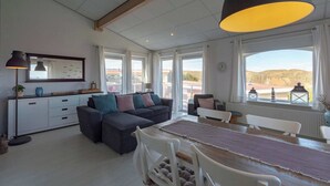 A spacious living area with a sofa, dining table, and chairs, large windows with curtains, and a ceiling with exposed beams. - Beachside Home With Balcony (Julianadorp)