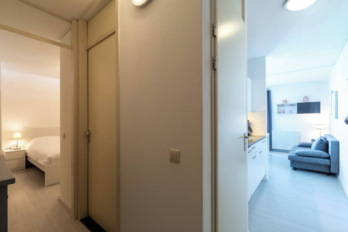 Ένα δωμάτιο ξενοδοχείου με ένα κρεβάτι, ένα κομοδίνο και ένα φωτιστικό. Υπάρχει μια πόρτα που οδηγεί σε ένα άλλο δωμάτιο.