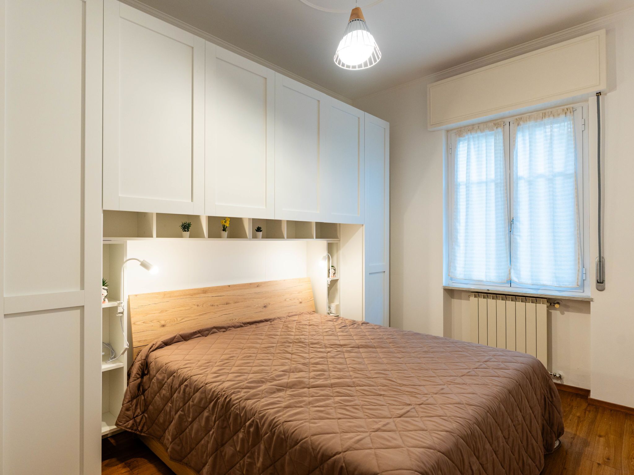 Una camera da letto con un letto, mobili bianchi, una finestra con tende e un termosifone.