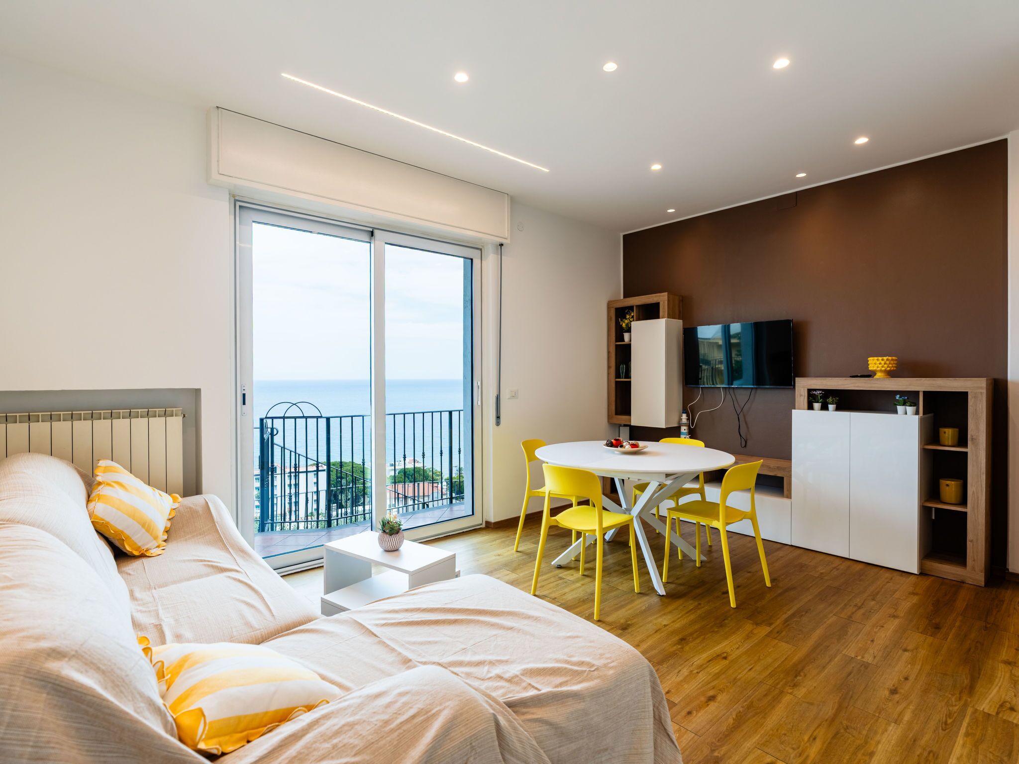 A modern bedroom with a large bed, a balcony with a view, a dining area with yellow chairs, and a TV on the wall.