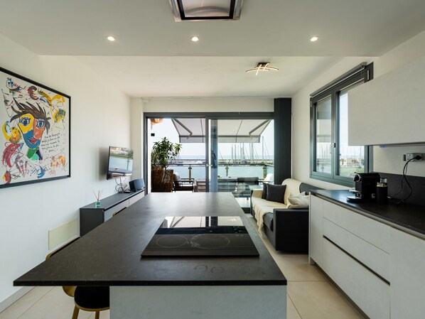 A modern kitchen with a large island, a dining area with a view of the marina, and a living area with a sofa and coffee machine.