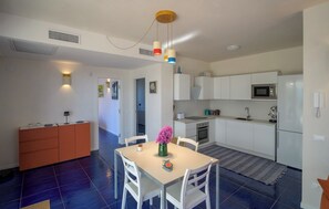 A modern kitchen with white cabinets, a microwave, and a refrigerator. There is a dining area with a table and chairs, and a vase with flowers. - Ginepro - Shelter by the sea in Apulia | A few steps from the beach (Puglia)