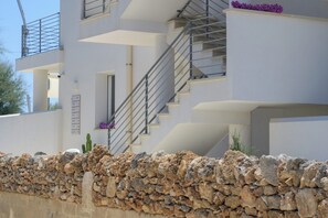 A modern multi-story building with a stone wall foundation, a staircase, and a balcony with purple flowers. - Ginepro - Shelter by the sea in Apulia | A few steps from the beach (Puglia)
