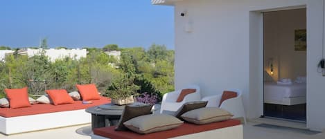A modern outdoor seating area with white sofas and red cushions, a small table with a vase, and a view of the surrounding greenery and buildings.