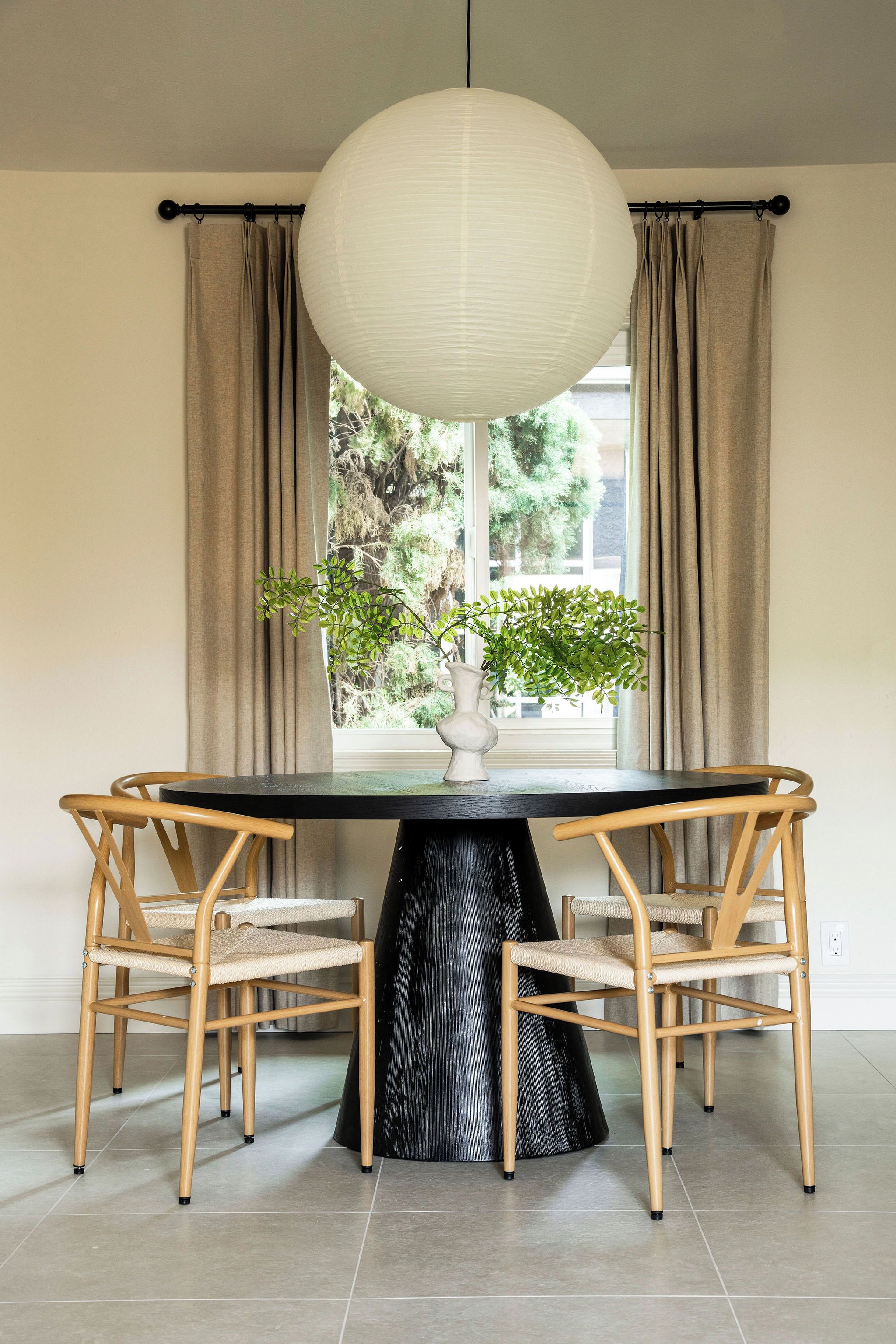 A dining table with a black base, surrounded by four wooden chairs with woven seats, a white vase with green plants, and a large white pendant light.
