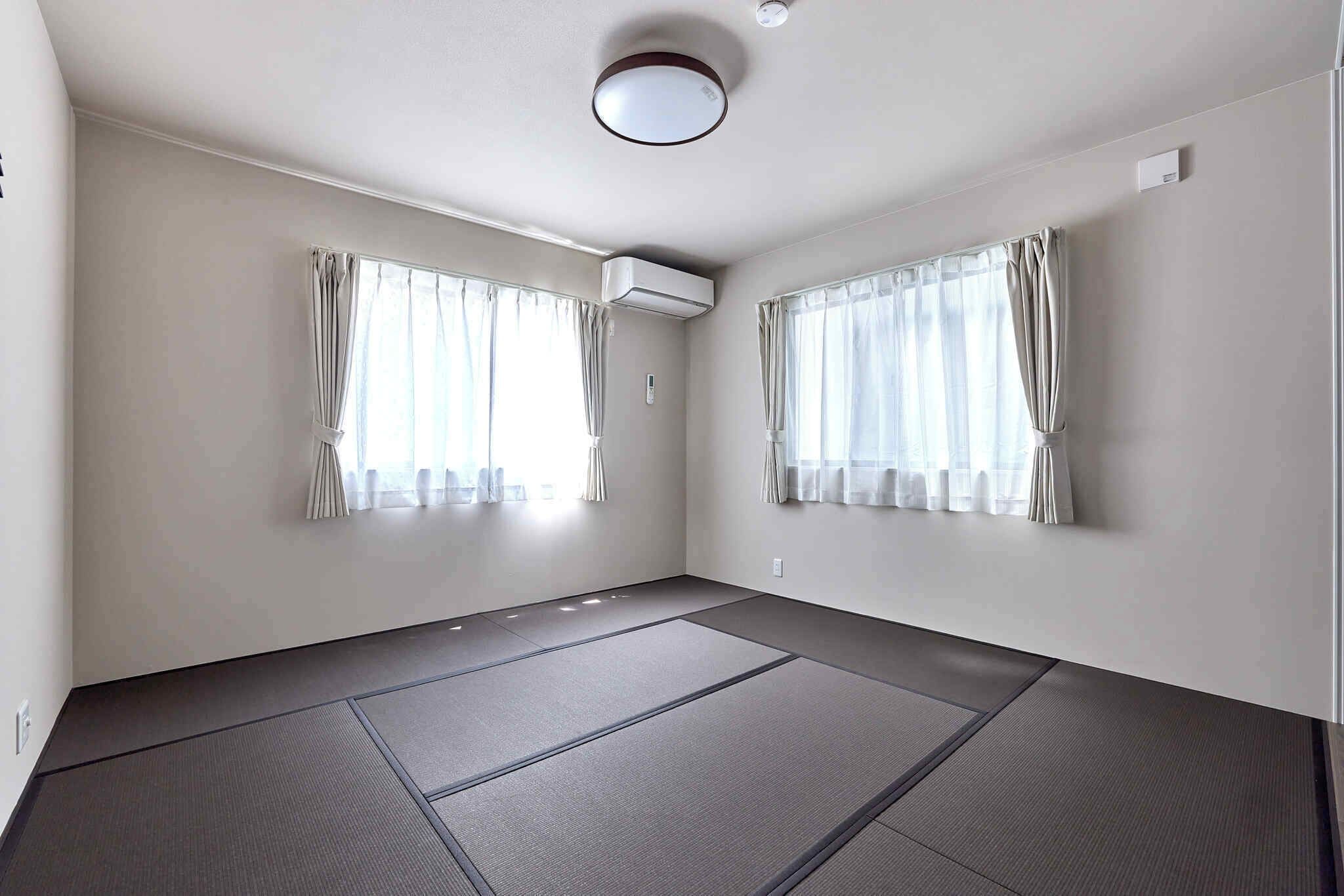 A room with tatami mats, two windows with curtains, an air conditioning unit, and a ceiling light.