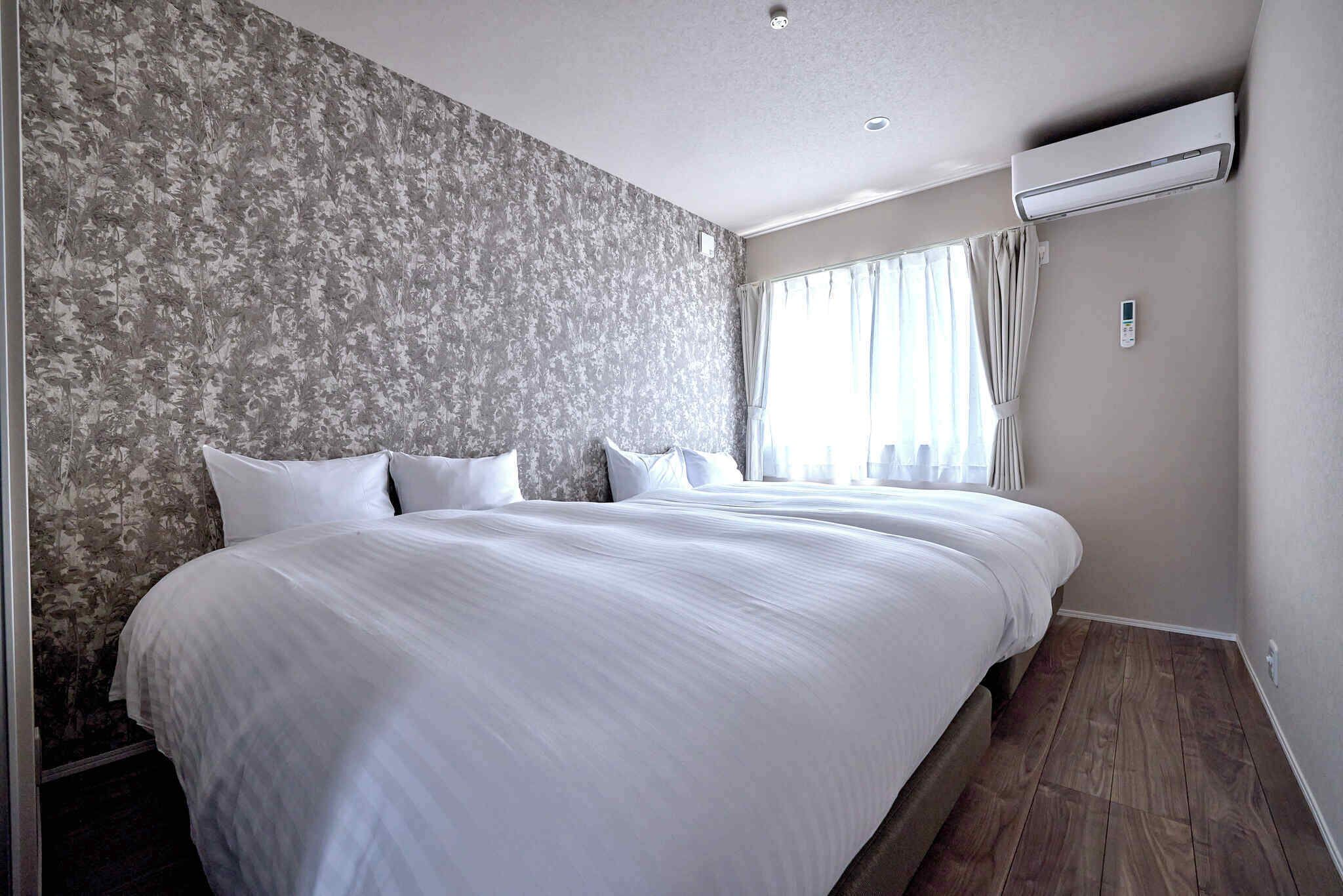 A hotel room with a large bed, floral wallpaper, an air conditioning unit, and a window with curtains.