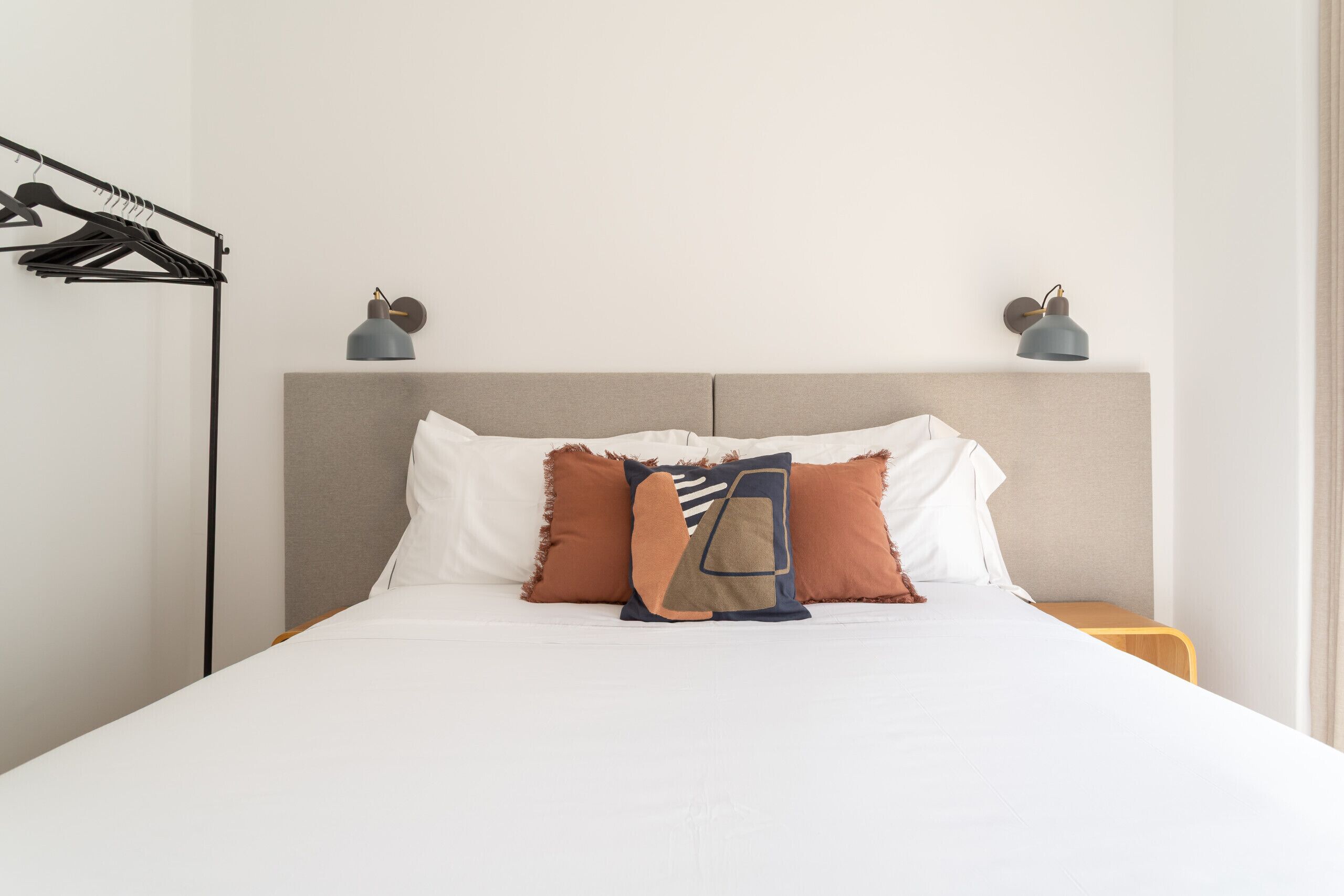 Una cama bien hecha con edredón blanco, dos almohadas y una manta decorativa. Hay una mesita de noche con una lámpara y una repisa en la pared.