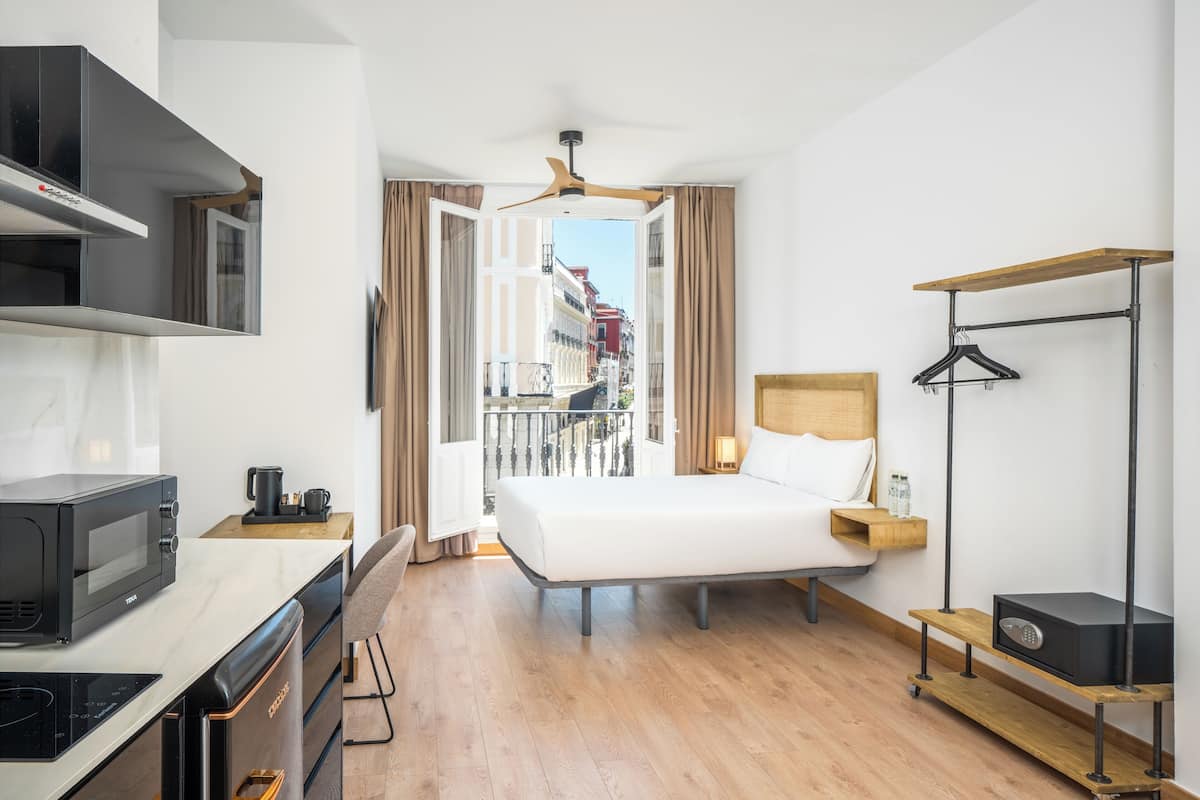 A modern hotel room with a bed, a desk with a microwave, a coffee maker, a hanging coat rack, and a view of the city through the open window.