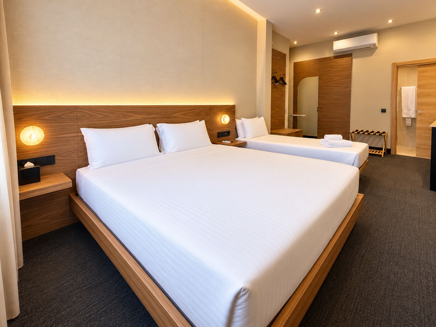 A hotel room with two beds, a wooden headboard, a bedside table with a lamp, a mirror, and an air conditioning unit.