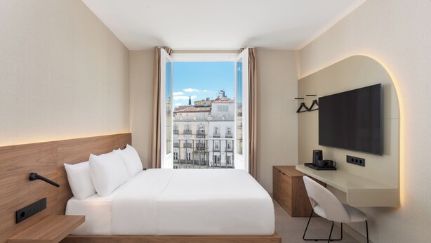 Una habitación de hotel moderna con una cama grande, un escritorio, un televisor de pantalla plana y vistas a la ciudad a través de un ventanal amplio.