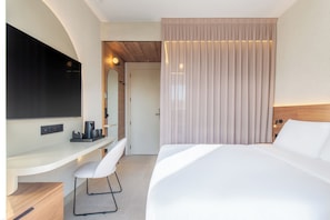 Ein modernes Hotelzimmer mit einem großen Bett, einem Schreibtisch mit Stuhl, einem Flachbildfernseher und einem großen Fenster mit Vorhängen.