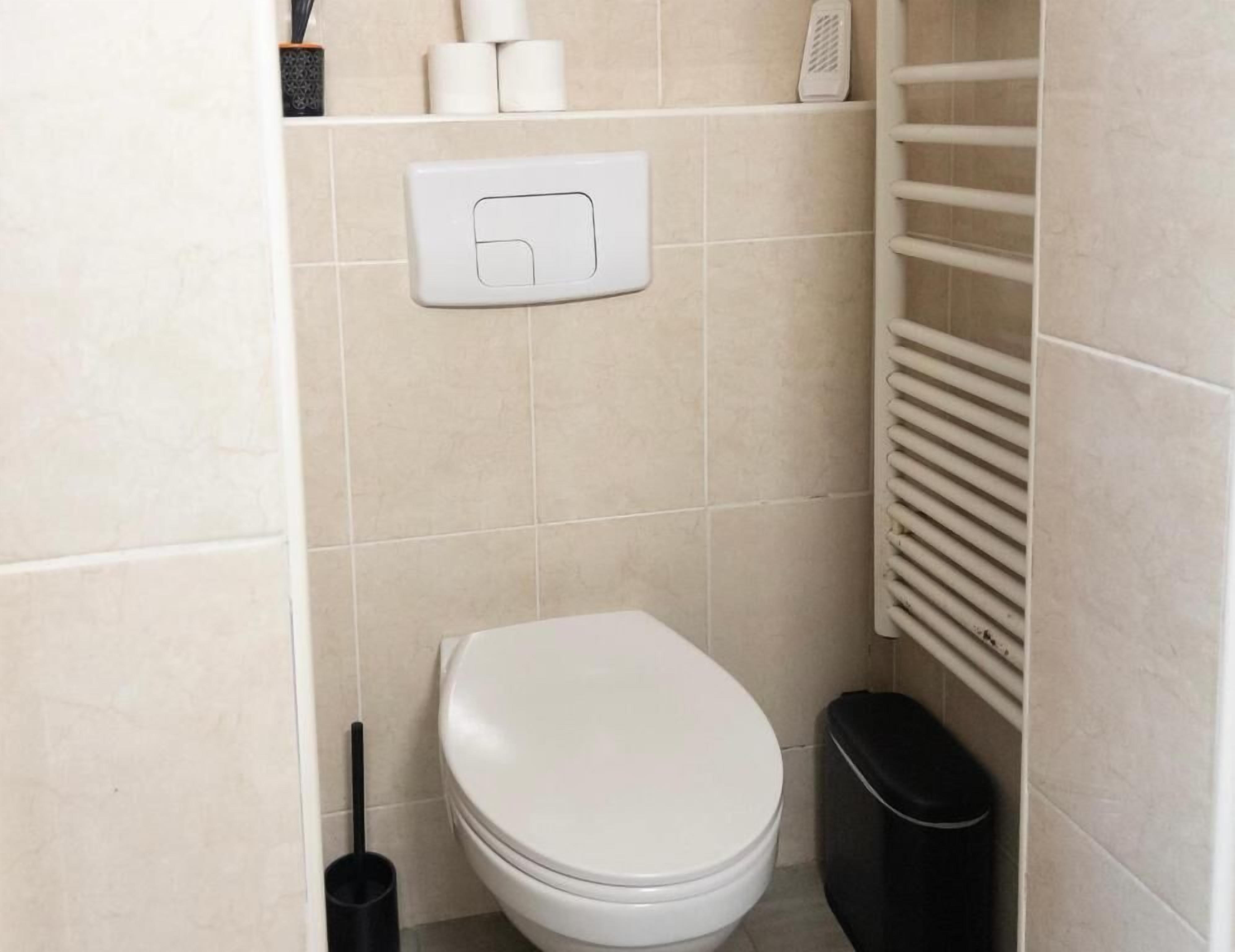 Egy kis, tiszta fürdőszoba, amelyben található egy WC, egy szemeteskosár és egy WC-kefe.