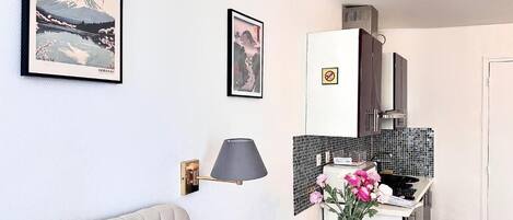 A compact kitchen with a dining area, featuring a table with a vase of flowers, a lamp, and framed artwork on the walls.