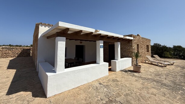 Un pequeño edificio blanco con un muro de piedra de fondo, un espacio de estar cubierto y una planta en maceta.