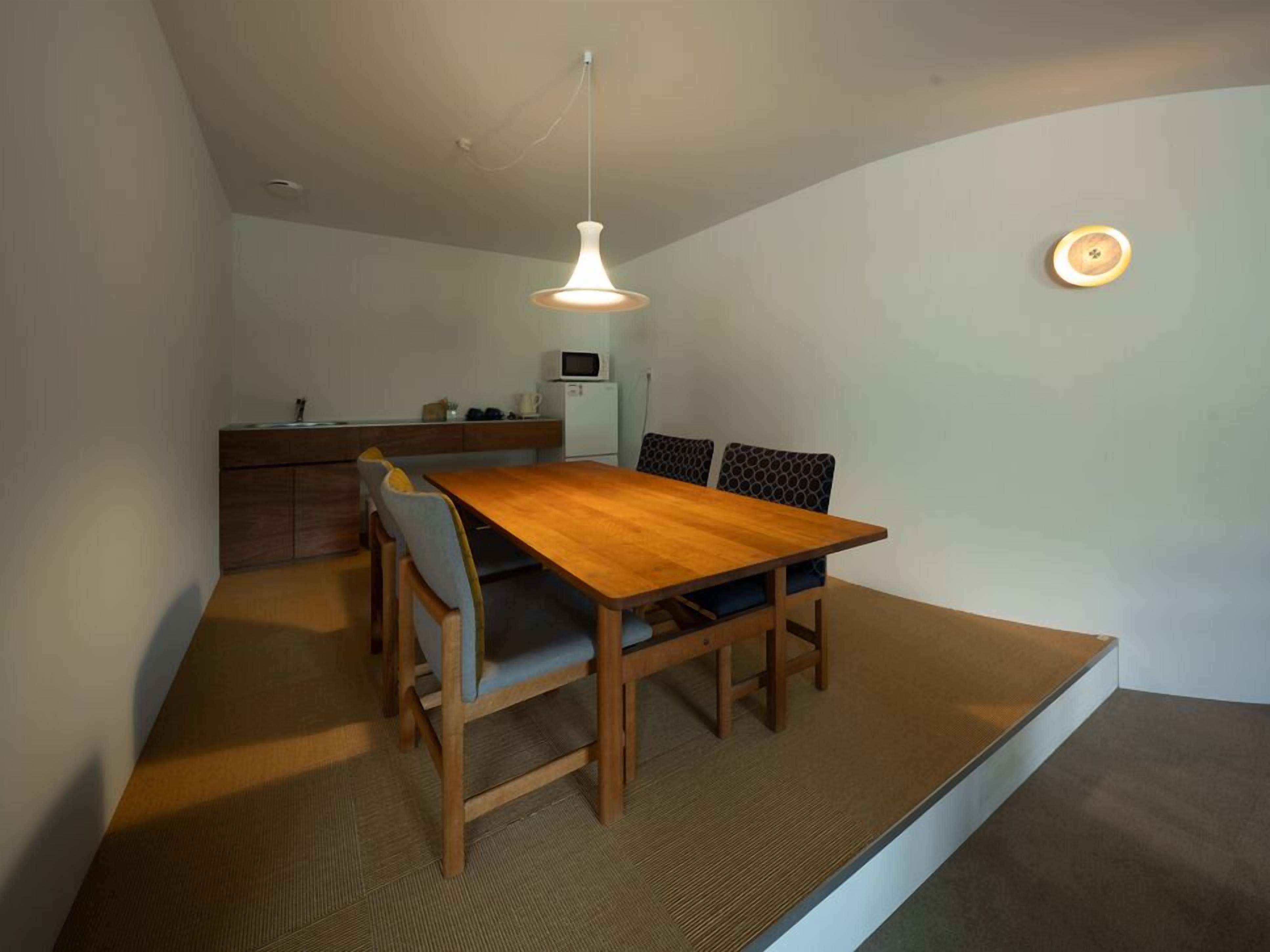 A compact dining area with a wooden table and chairs, a hanging light fixture, and a kitchenette in the background.
