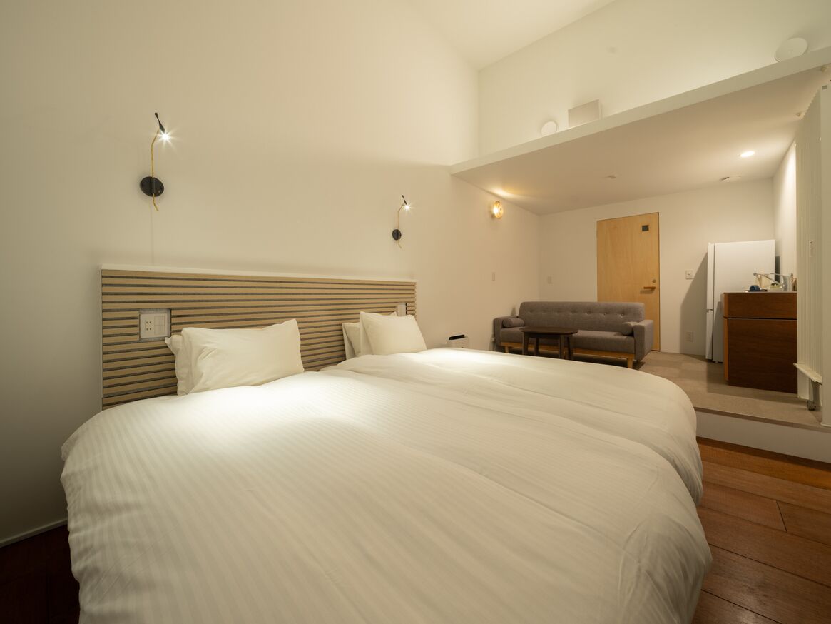 Une chambre à coucher avec un grand lit, une tête de lit en bois, un fauteuil gris, un bureau avec une lampe et une porte donnant sur une autre pièce.