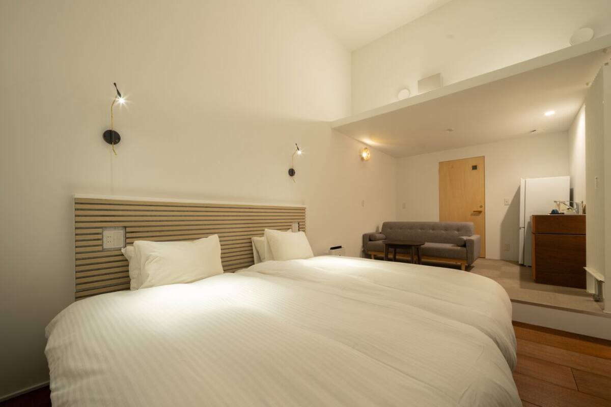 Quarto com cama grande, cabeceira de madeira, poltrona cinzenta, escrivaninha com abajur e porta para outro cômodo.