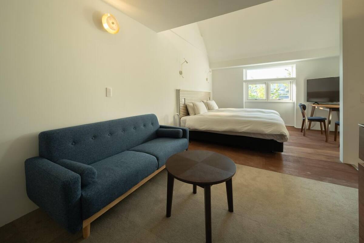 Quarto com cama, sofá azul, mesinha, televisão e janela.