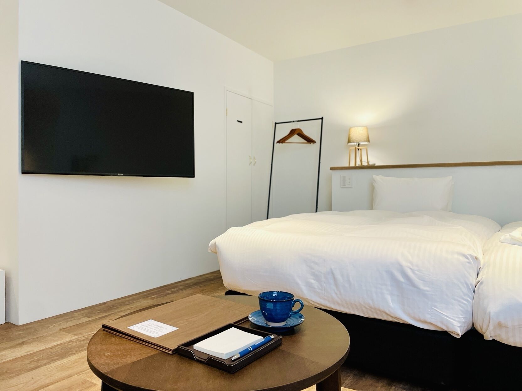Une chambre d’hôtel avec un lit, un téléviseur fixé au mur, une table de chevet avec une tasse et une assiette, et une lampe fixée au mur.