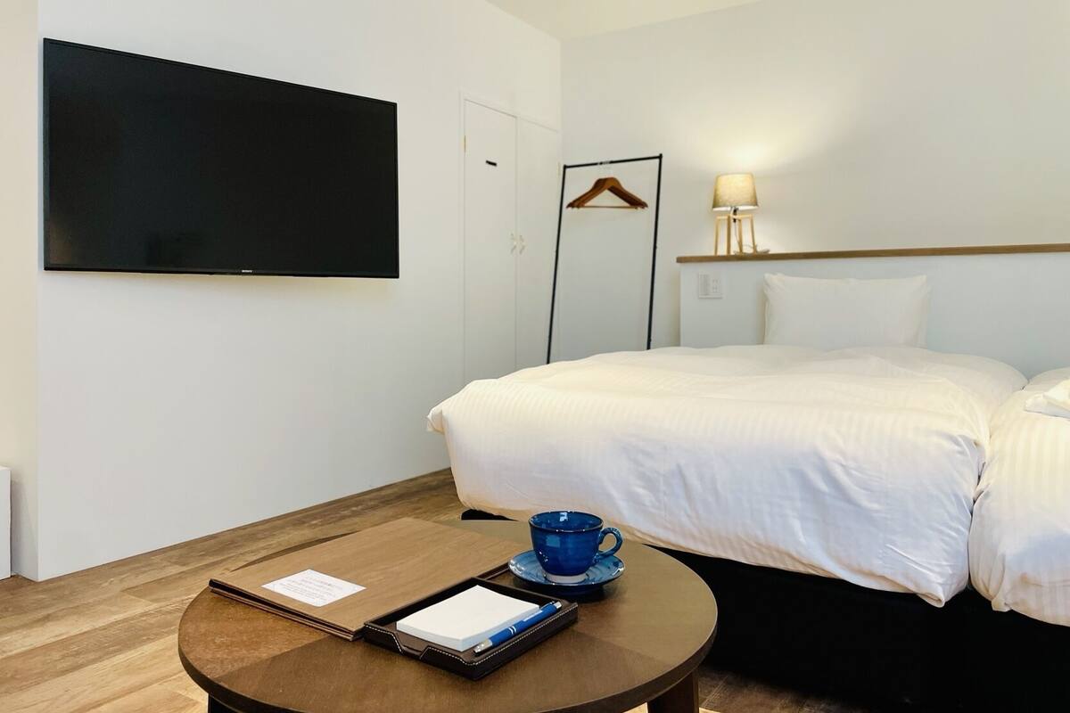 Quarto de hotel com cama, televisão na parede, mesa de cabeceira com uma chávena e um tabuleiro, e um candeeiro na parede.