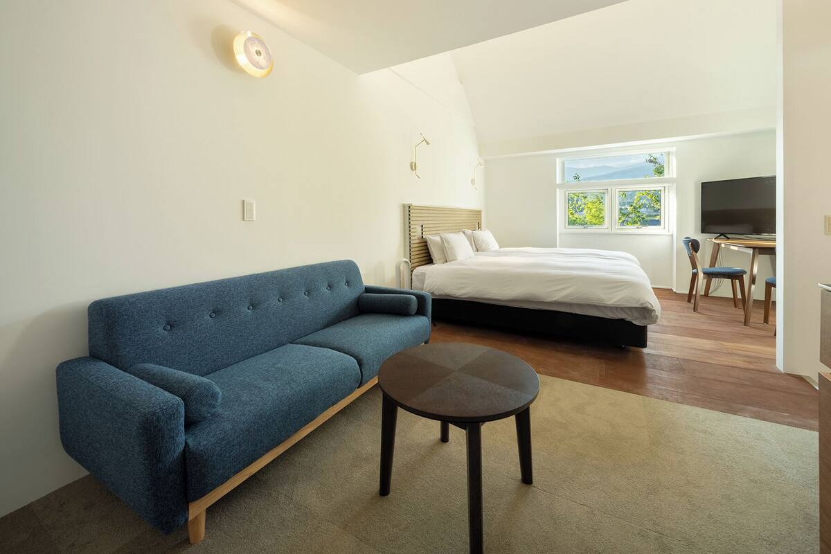 Quarto moderno com sofá azul, cama com roupa de cama branca, mesa redonda pequena e TV num canto.