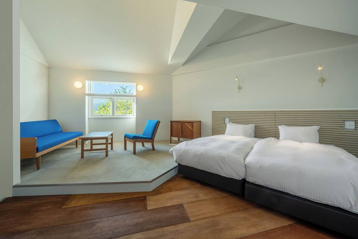 Quarto com uma cama grande, um sofá, uma cadeira e uma mesa pequena. Há uma janela com vista para o verde.