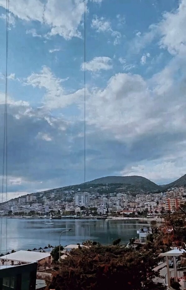 A coastal cityscape with buildings, a body of water, and a hill in the background. - Hotel MS (Sarandë)