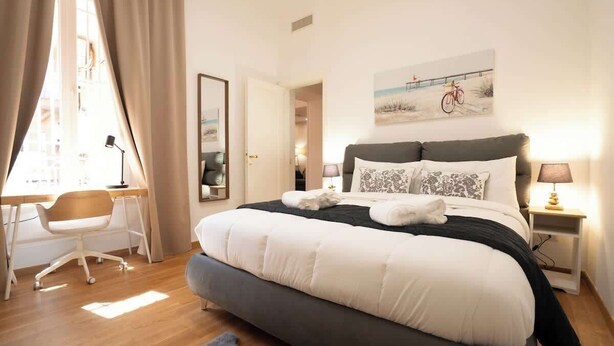 Un dormitorio con una cama grande, un escritorio y una silla. Hay un cuadro en la pared y una lámpara en la mesita de noche.