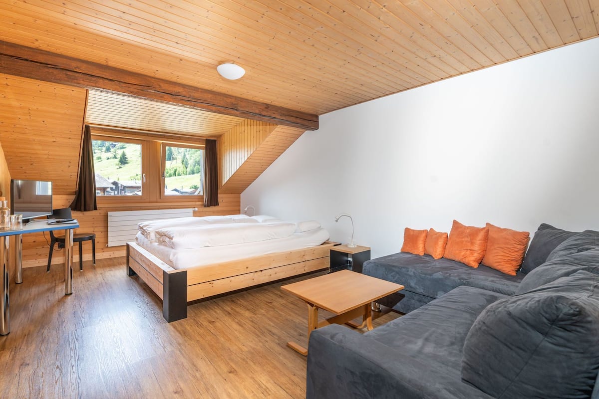 A modern bedroom with a wooden ceiling, a bed, a desk, a sofa, and a window with a view of greenery.