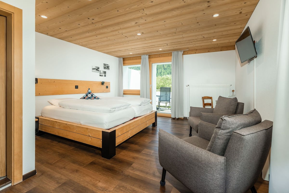 A modern bedroom with a wooden ceiling, a large bed, a grey sofa, and a TV on the wall.