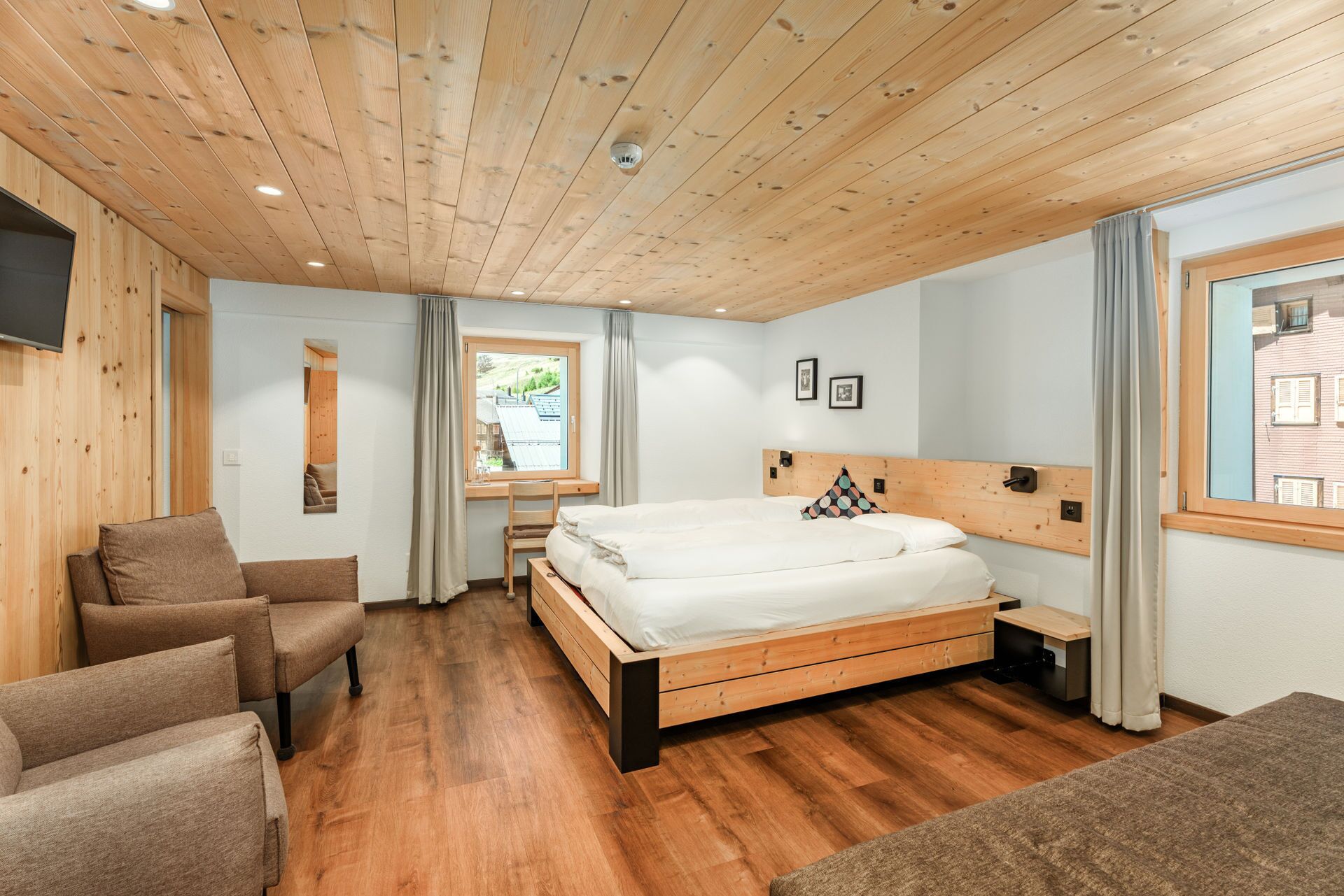 A bedroom with a wooden ceiling, a bed with a wooden headboard, a desk, a chair, and a window with a view of a building.