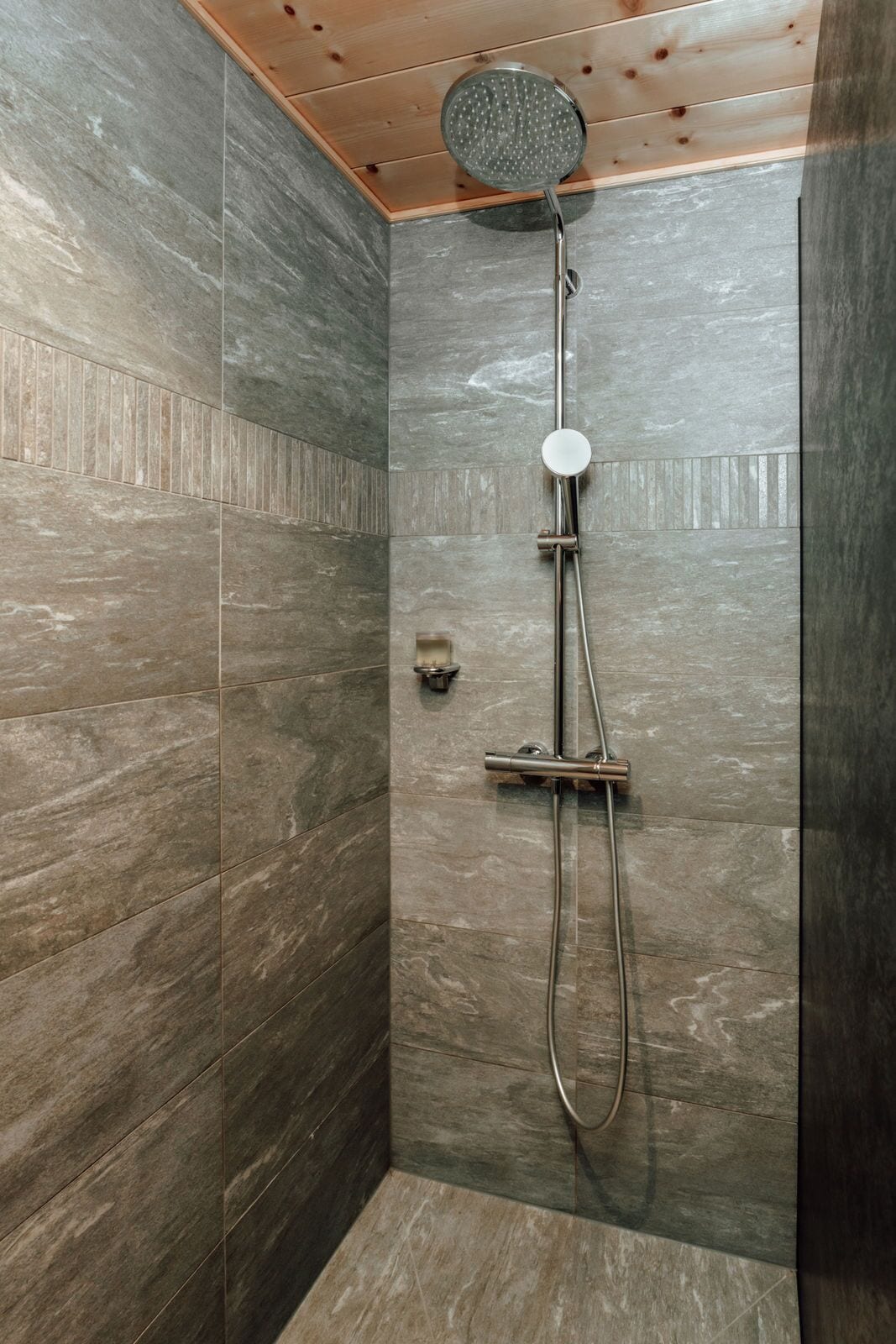 A shower with a handheld showerhead and a wall-mounted faucet, set in a room with wooden ceiling and stone-tiled walls.