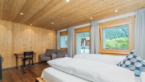 A bedroom with a wooden ceiling, a bed with white bedding, a desk, a chair, and a view of a forest through large windows. - Hotel Furka (Obergoms)