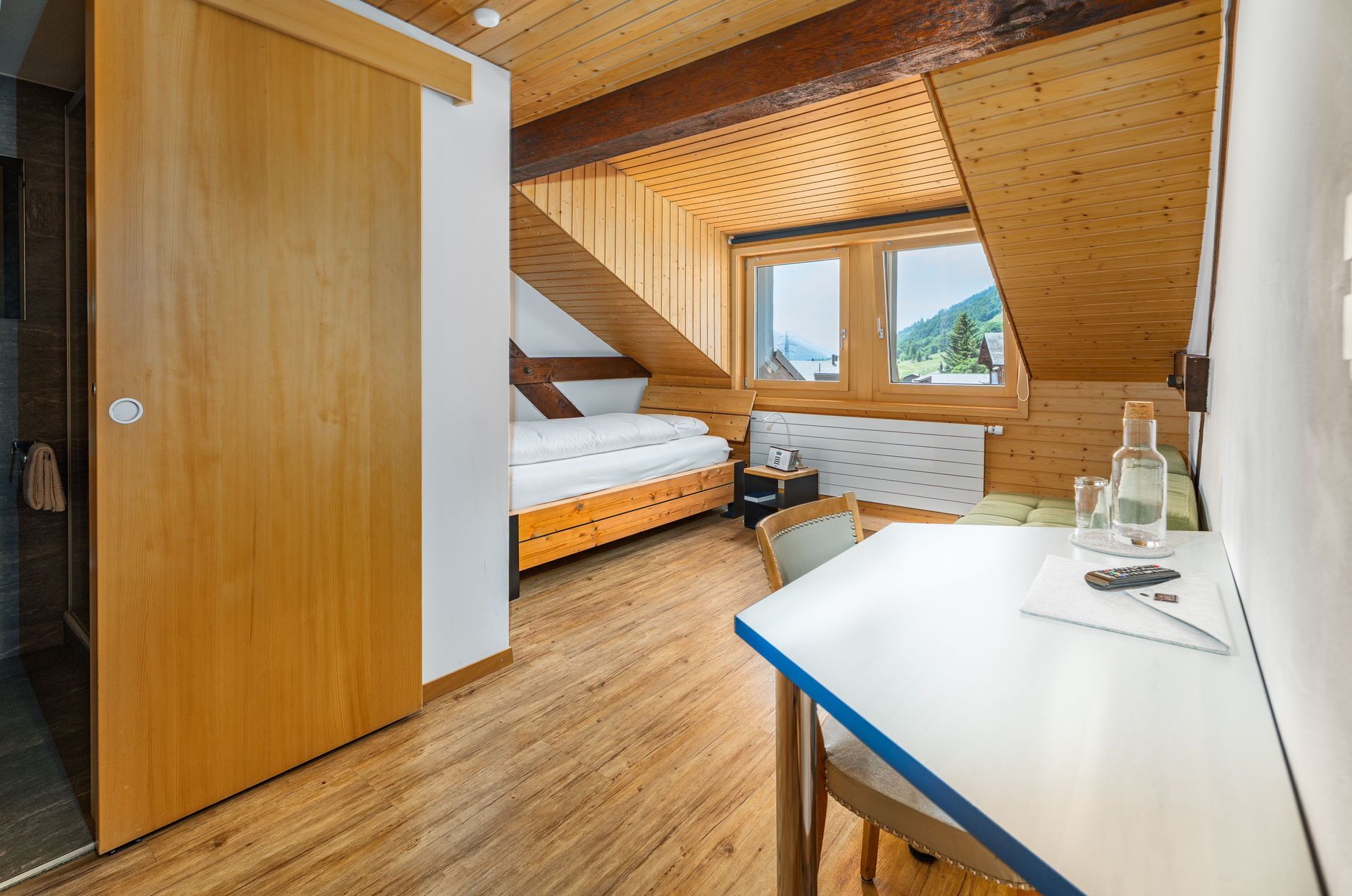 A compact bedroom with a wooden ceiling, a bed, a small table, and a window with a view of the outdoors.