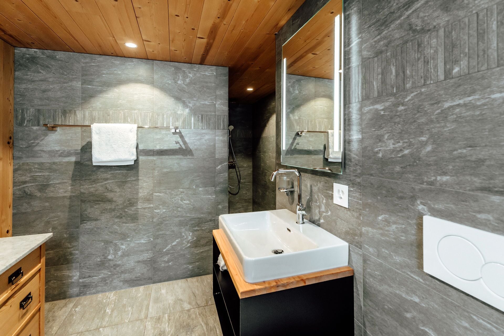 A modern bathroom with a wooden ceiling, grey marble walls, a white sink, and a large mirror.