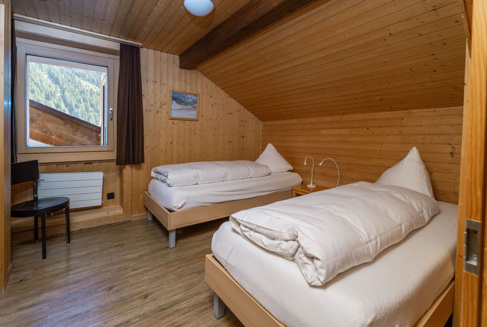 A room with two beds, a chair, a radiator, and a window with a view of trees.