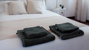A neatly made bed with a beige bedspread and pillows, a bedside table with a lamp and a small plant, and a stack of folded towels on the bed.