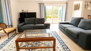 A modern living room with a glass coffee table, two grey sofas, a wooden chair, and a black entertainment unit.