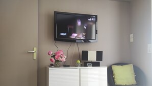A living room with a TV mounted on the wall, a white cabinet, a black chair with a cushion, and a vase with pink flowers. - Rent T2 a St Aygulf (St Aygulf)