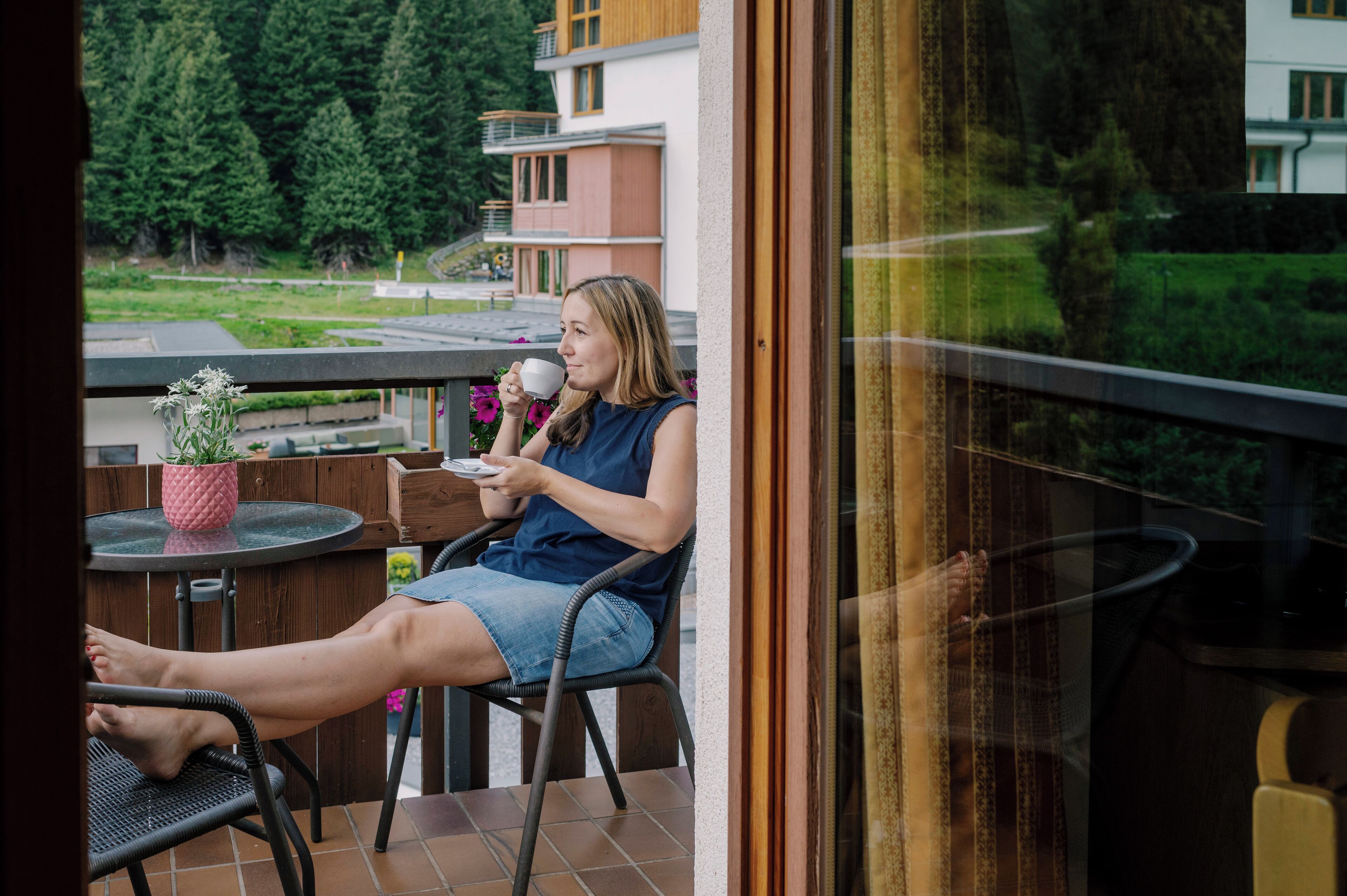 Una persona sentada en una silla de balcón, sosteniendo una taza y un plato, con vistas a un paisaje verde y edificios al fondo.
